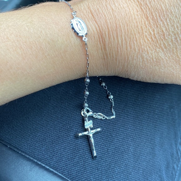 Silver rosary bracelet with a cross pendant on a wrist against a dark blue fabric background availabe on TX.ORO website.
