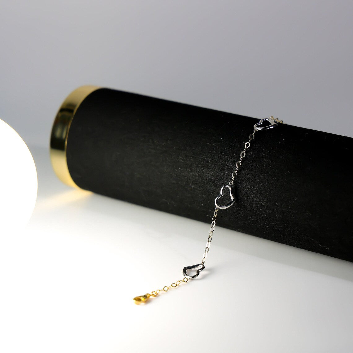 Delicate silver bracelet with heart-shaped charms on a black cylindrical stand against a gray background available on the TX.ORO website.