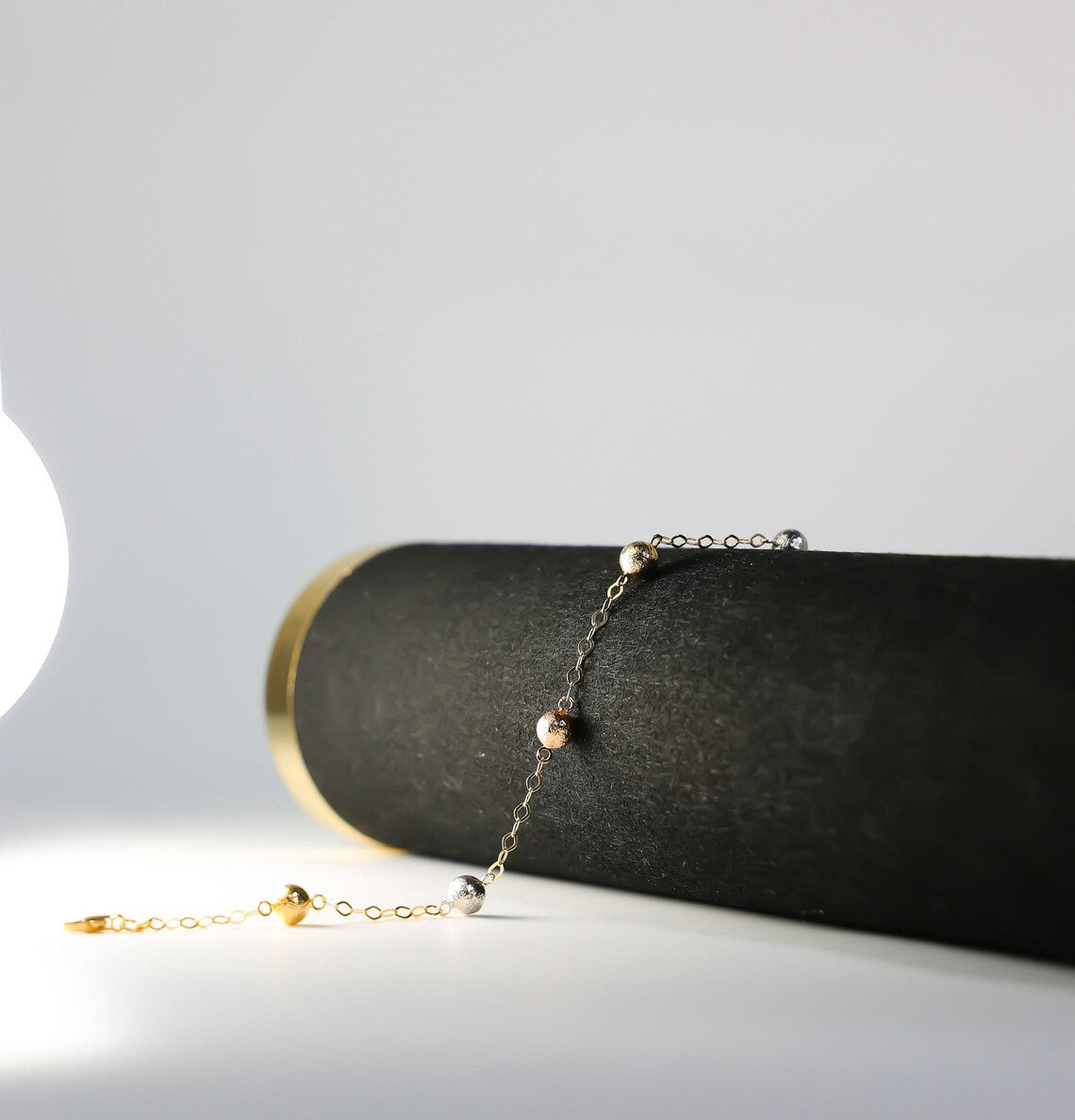 Gold bracelet with small beads on a black cylindrical object against a light gray background available on the TX.ORO website.