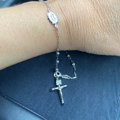 Silver rosary bracelet with a cross pendant on a wrist against a dark blue fabric background availabe on TX.ORO website.