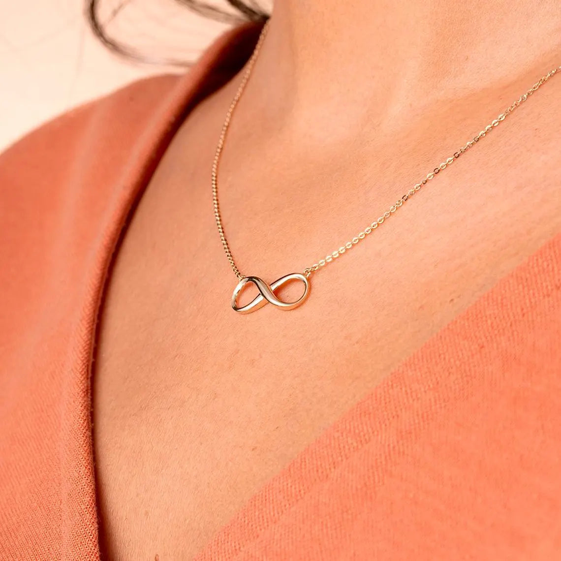 Gold infinity necklace on a person wearing a coral top available on the TX.ORO website.
