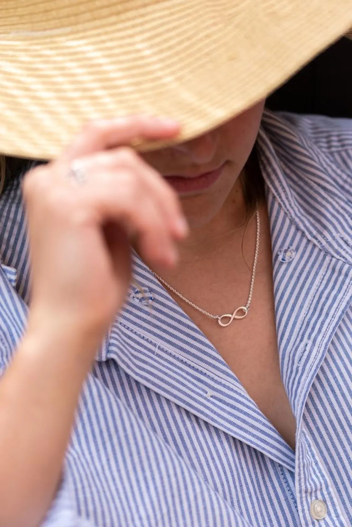 Person wearing a straw hat and a blue striped shirt with a necklace featuring an infinity symbol available on the TX.ORO website..