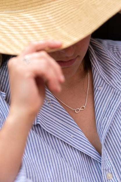 Person wearing a straw hat and a blue striped shirt with a necklace featuring an infinity symbol available on the TX.ORO website..