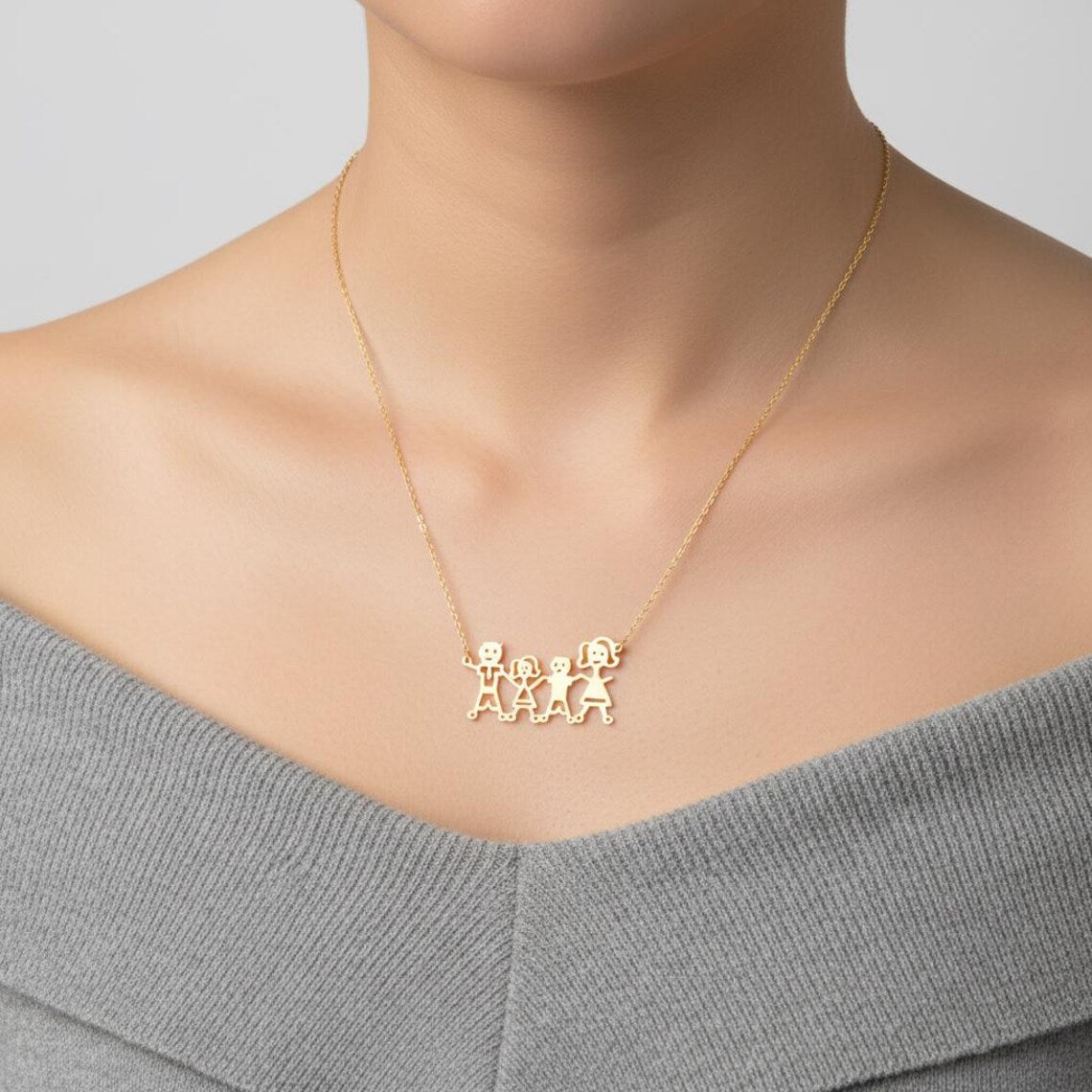 A close-up of a model wearing a delicate gold chain necklace with a pendant featuring three small connected figures, styled with a gray off-the-shoulder top.