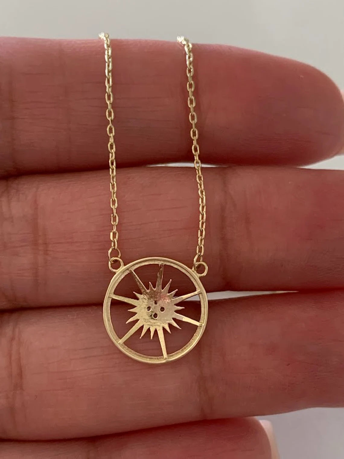 Gold necklace with a sun-shaped pendant held between fingers available on the TX.ORO website.