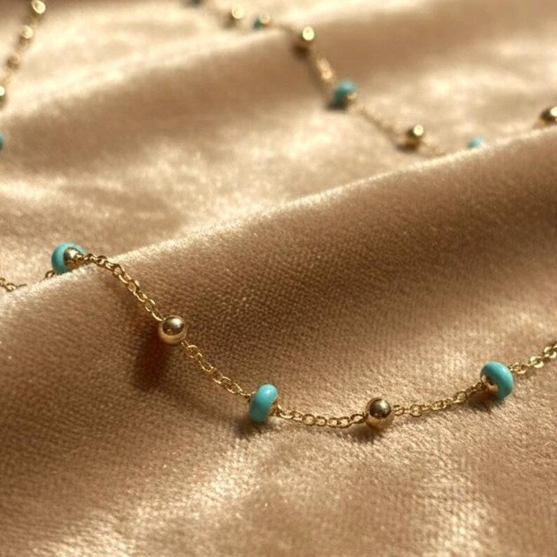 Gold necklace with turquoise beads on a beige fabric background available on the TX.ORO website.