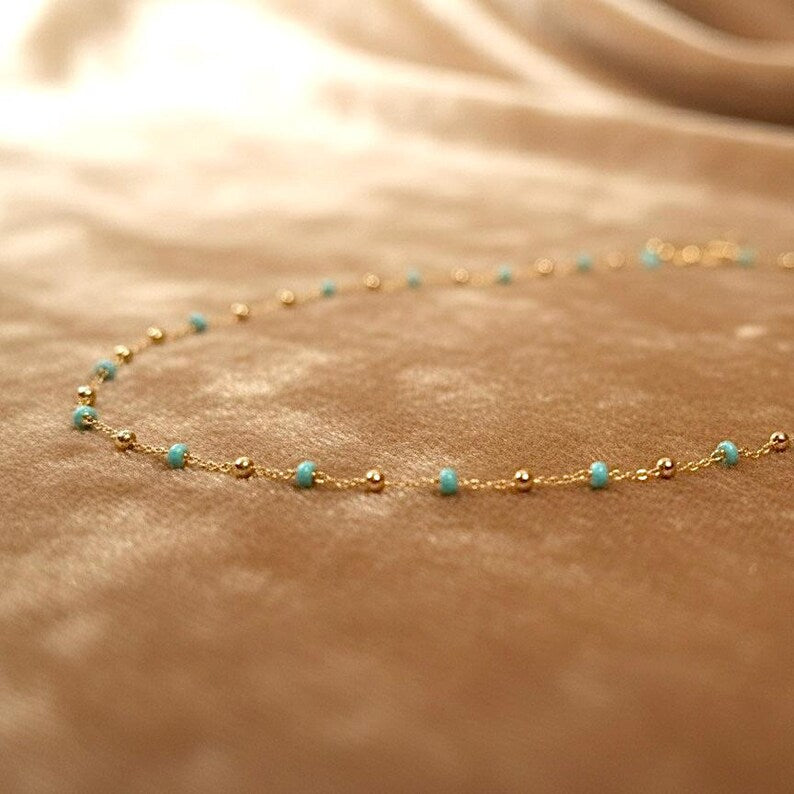 Gold and turquoise beaded necklace on a beige fabric background available on the TX.ORO website.