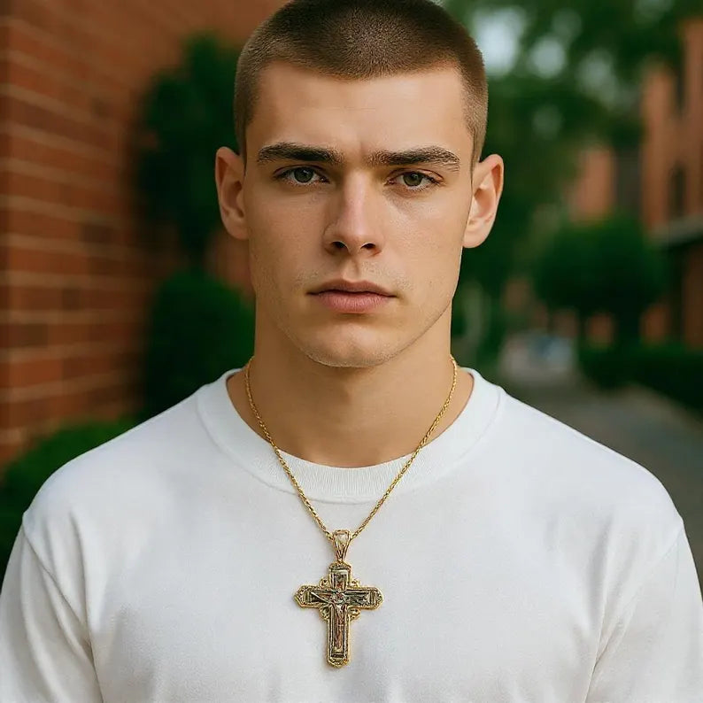 Man wearing a white shirt and gold necklace with a cross pendant in front of a blurred outdoor background.